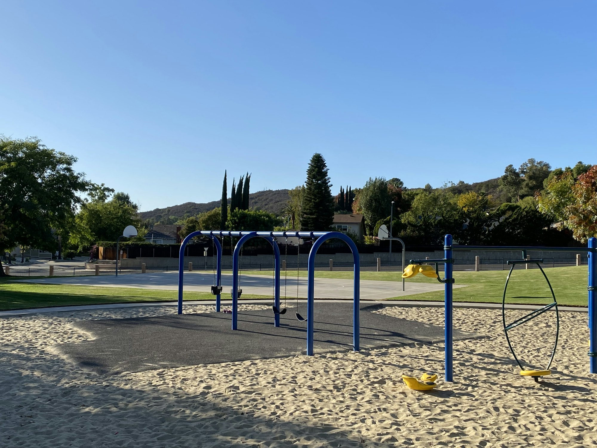 A playground with swings, basketball courts, and sand, surrounded by trees and hills in the background.