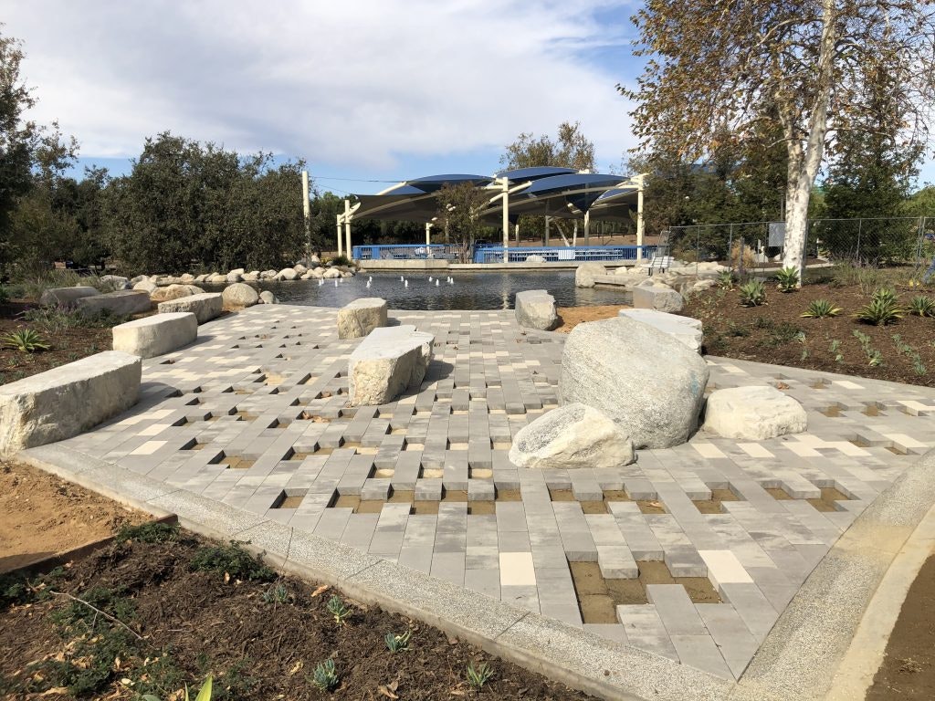A paved walkway with large stones leads to a water feature surrounded by trees and a covered seating area.
