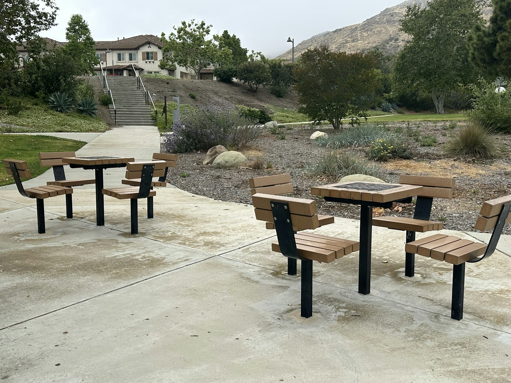 The image shows two sets of modern picnic tables and benches in a park, with a landscaped area and steps in the background.