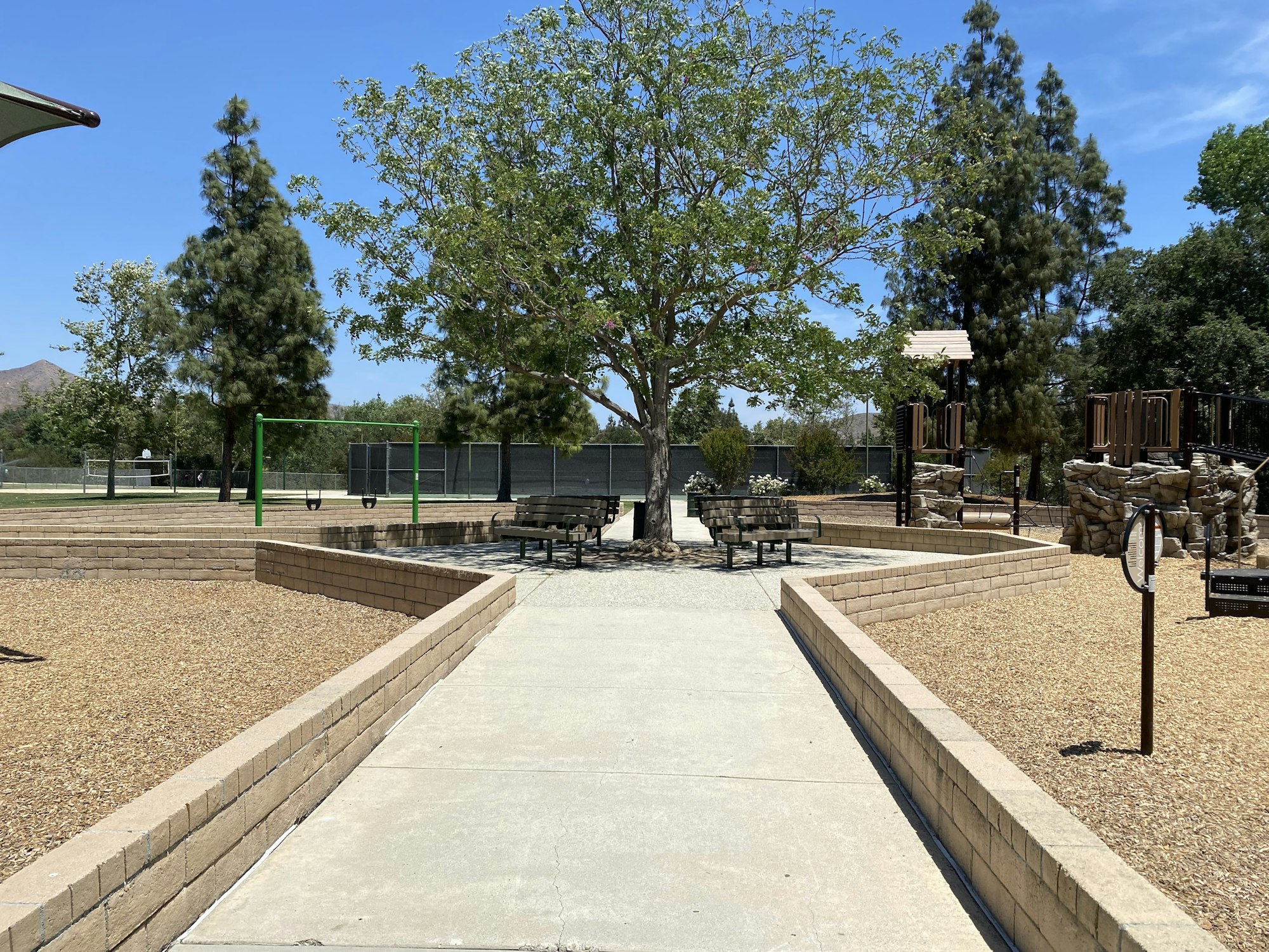 A park with a tree, benches, swings, playground equipment, and pathways under a clear blue sky.