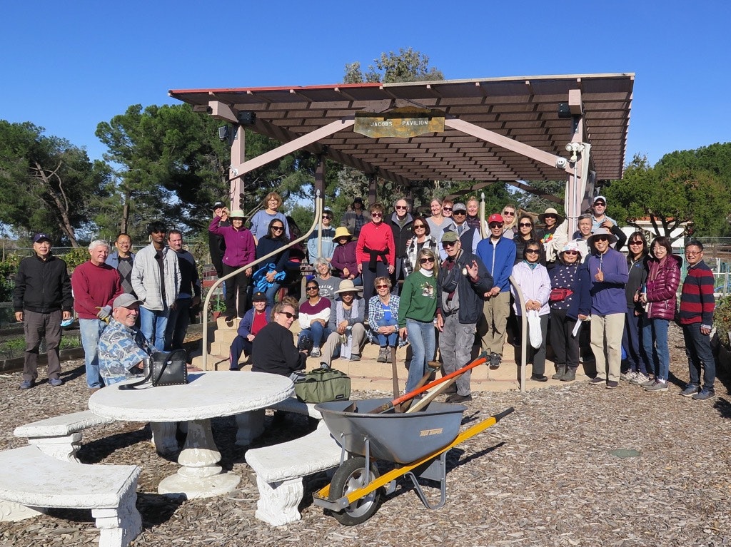 A group of people posing in front of a pavilion in a garden, with trees and a wheelbarrow in the foreground.