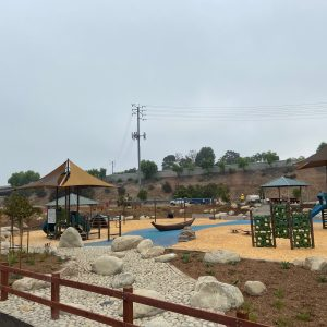 Outdoor playground with slides, climbing structures, shaded areas, and large rocks in a landscaped setting.