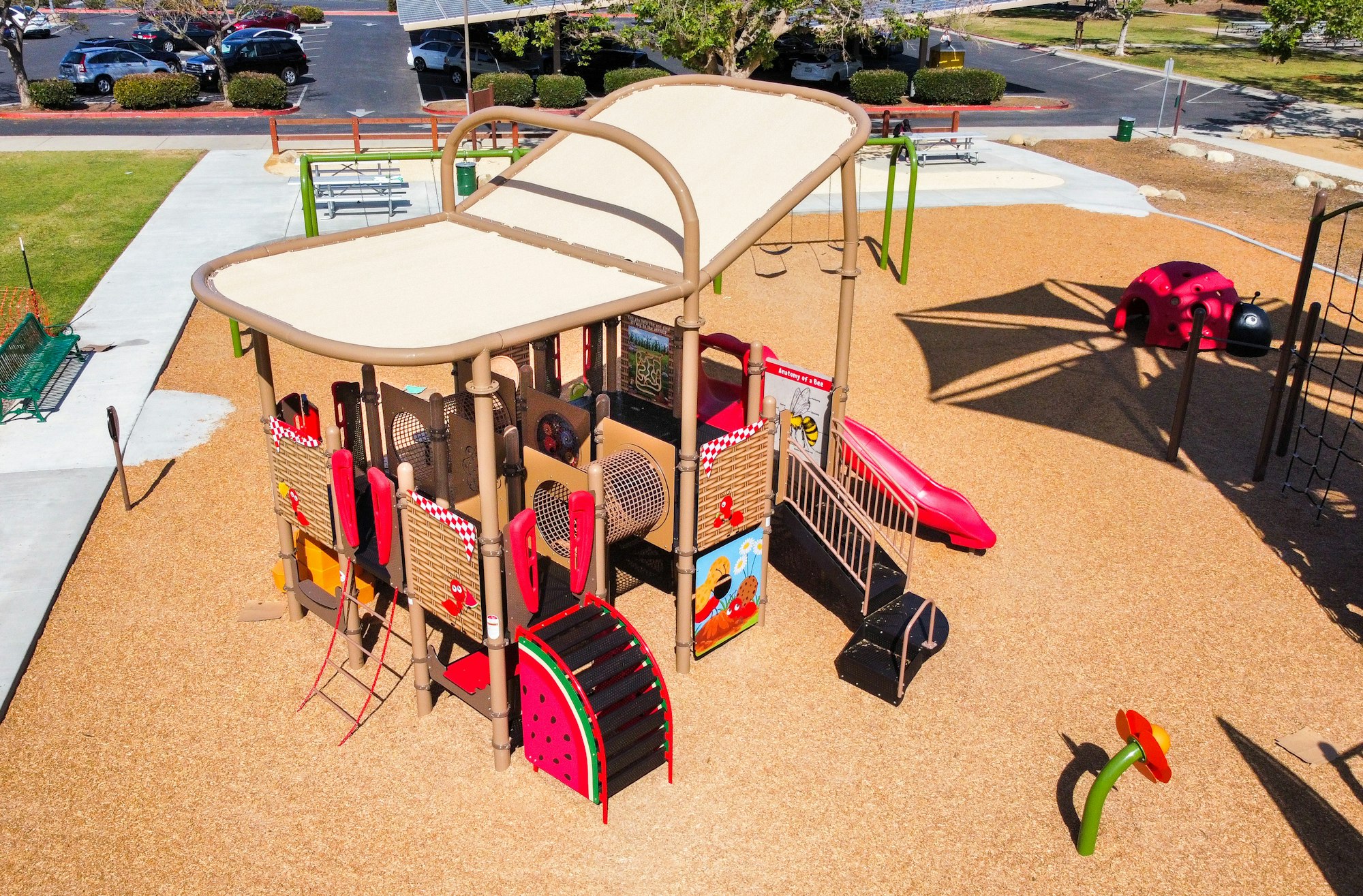 Playground with climbing structures, slides, swings, and a ladybug play equipment on tan ground, with parking area nearby.