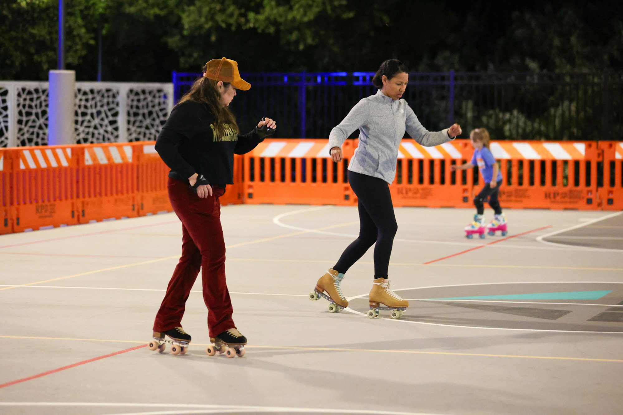 Two women are roller skating in an indoor rink, while a child skates in the background. Bright orange barriers are visible.