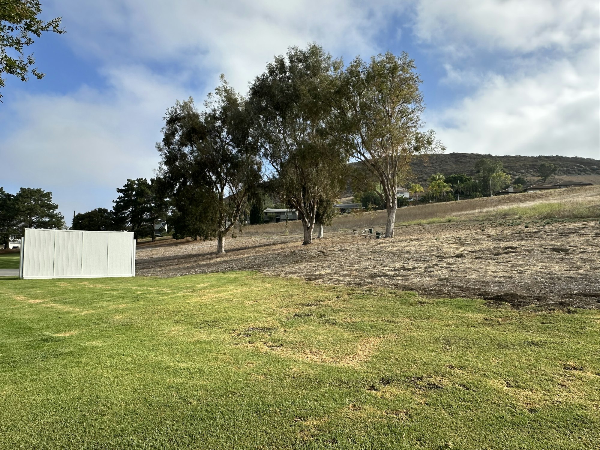 A grassy area with a white structure, trees, and a hillside under a partly cloudy sky.