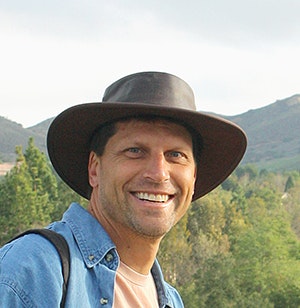 A person wearing a brown hat and blue shirt, smiling, with a hilly landscape in the background.