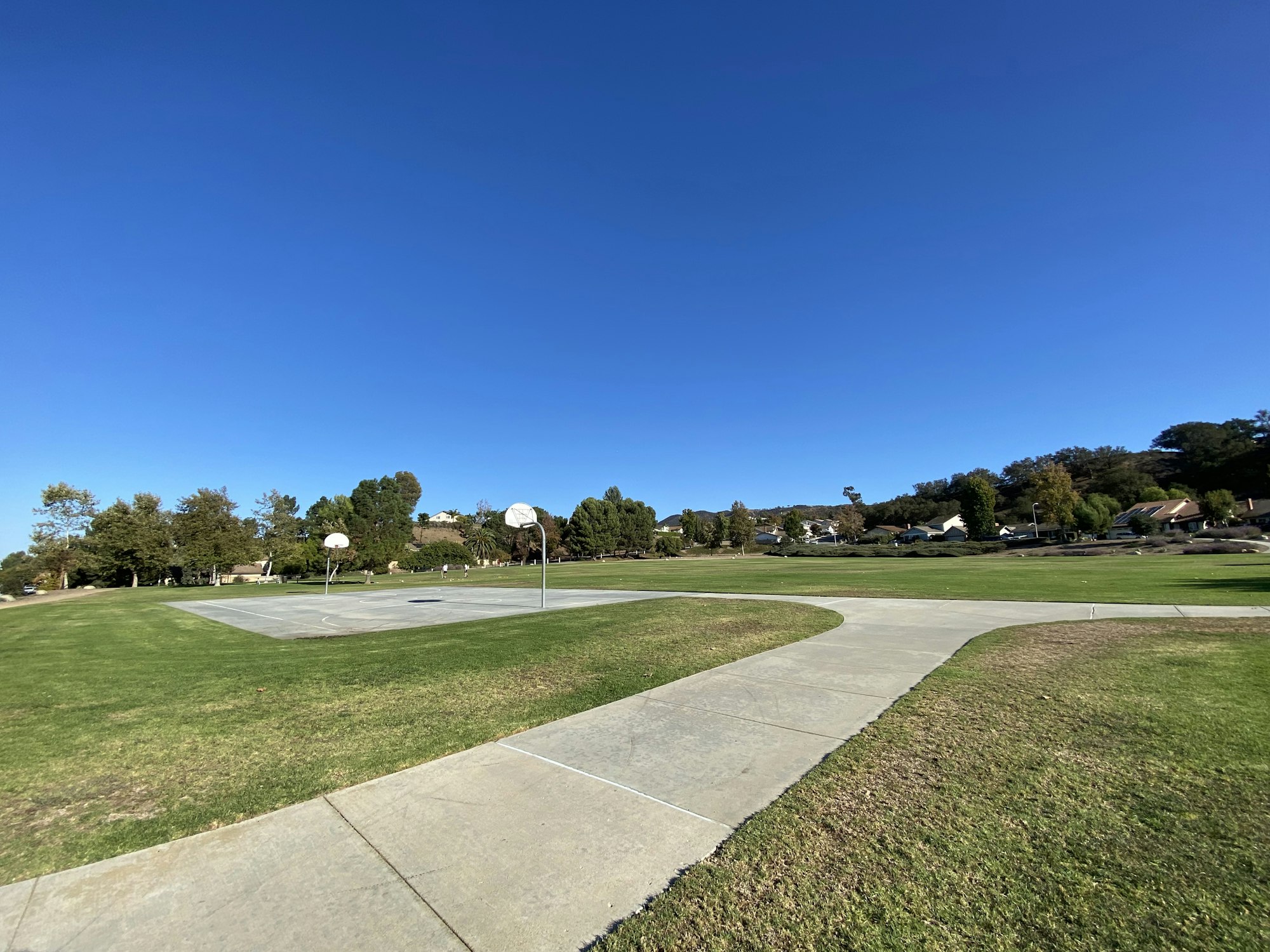 A park with a basketball court under a clear blue sky, surrounded by grass and trees.