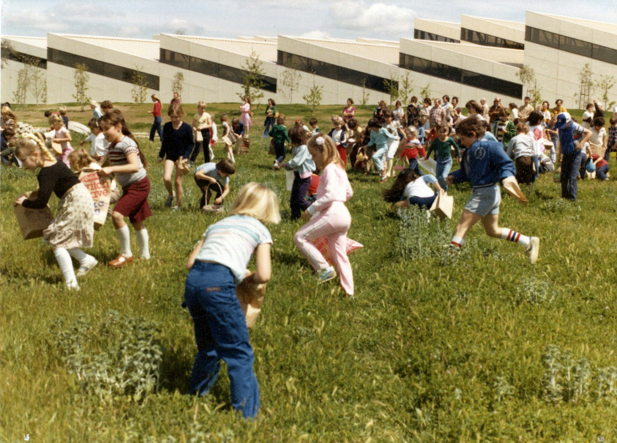 A large group of children playing outdoors on a grassy field, with some carrying bags, near a modern building.