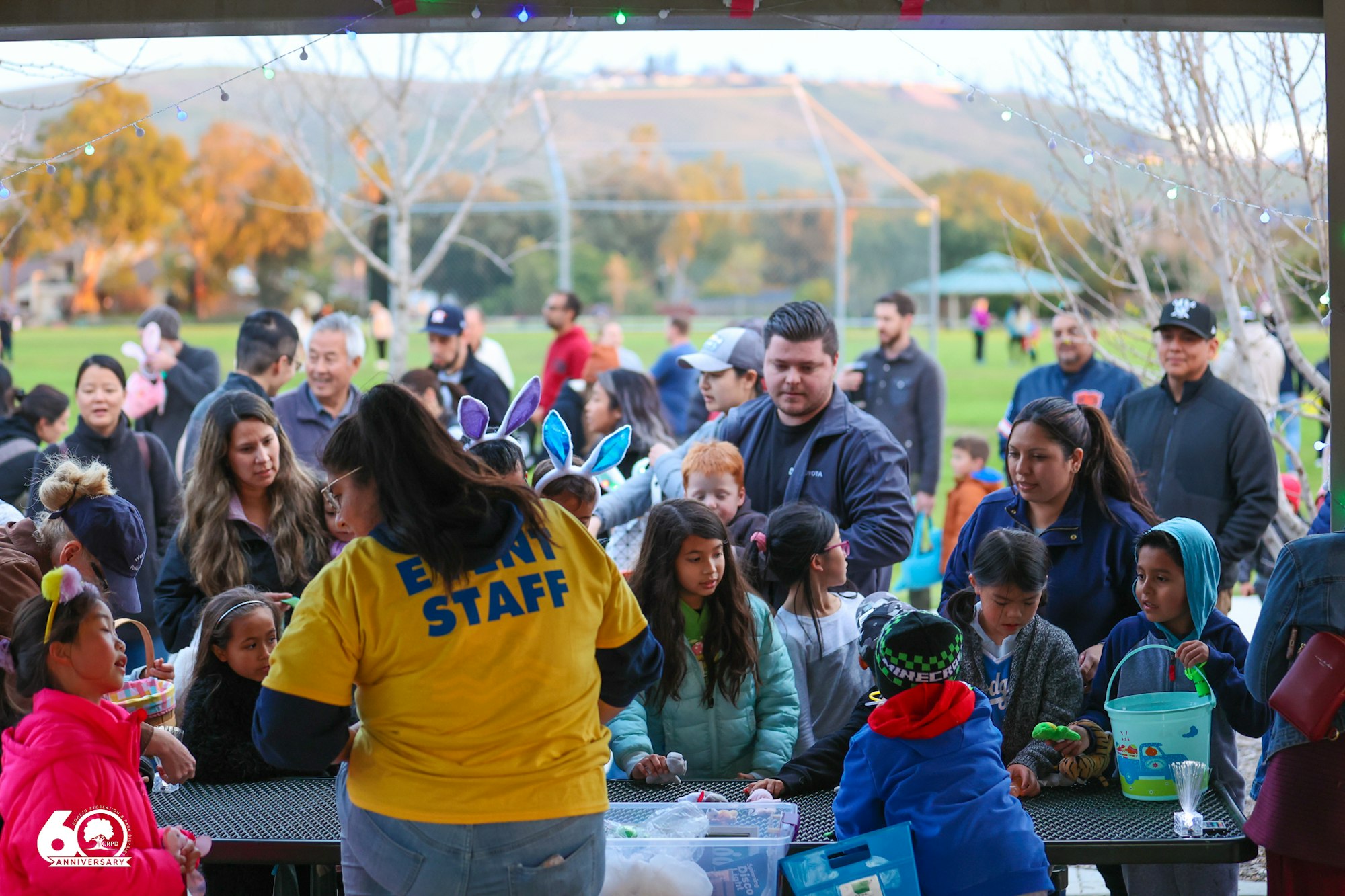 A community event with people gathering, event staff assisting, and children with baskets, possibly for a festive activity.