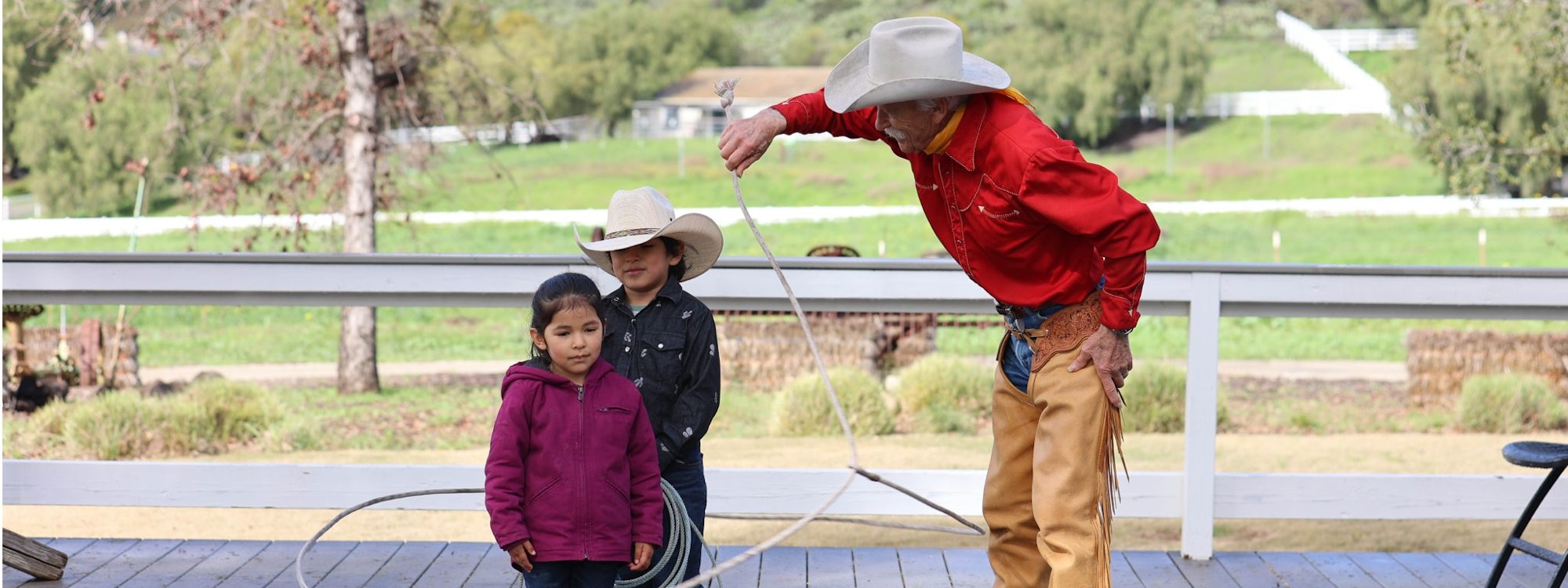 A cowboy teaches two children in cowboy hats how to use a lasso on a wooden platform.