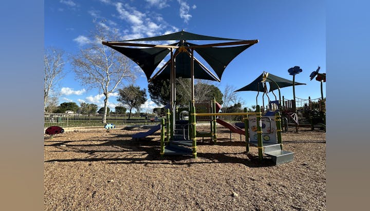 Playground with slides and shade structures under a clear blue sky. Trees and fences in the background.