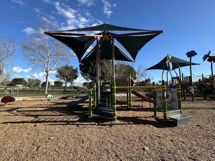 Playground with slides and shade structures under a clear blue sky. Trees and fences in the background.