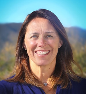 A person with long brown hair smiling, outdoors with a blurred background of hills under a blue sky.