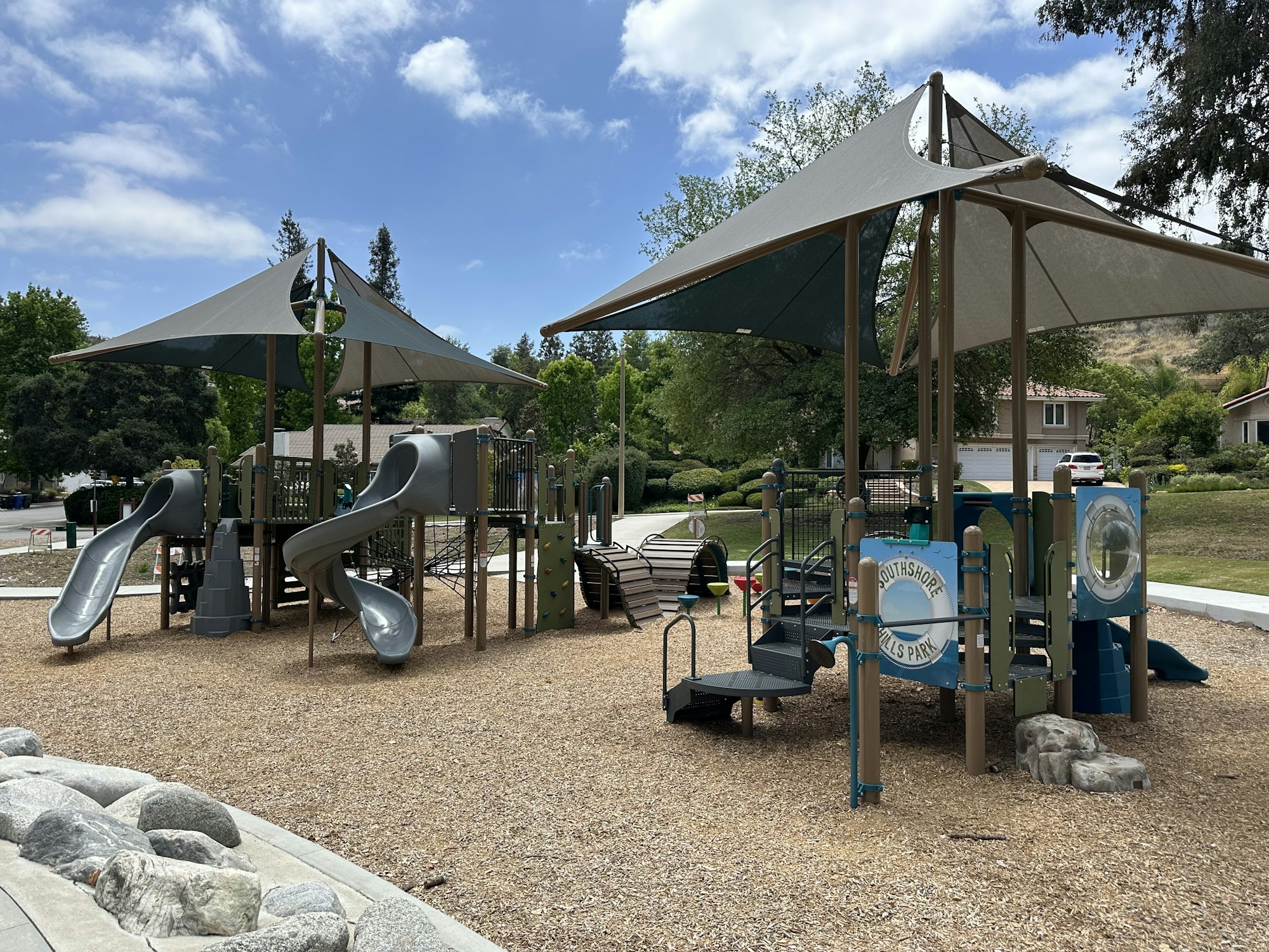 A playground with two slides, climbing structures, and shaded areas, set in a grassy environment on a sunny day.