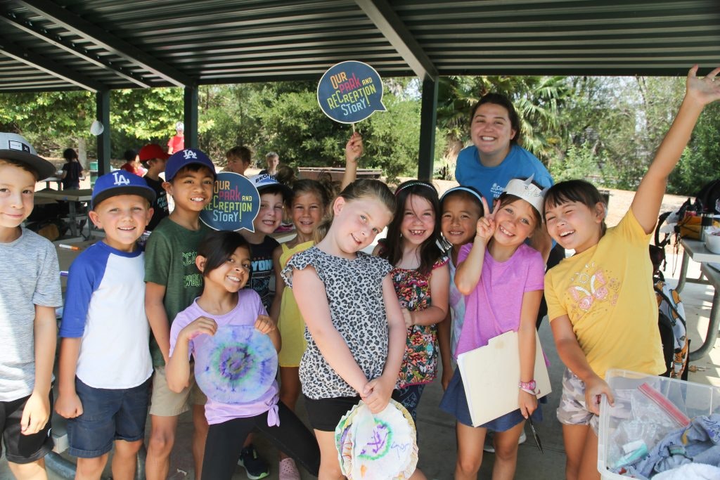 A group of smiling children and an adult pose under a shelter, holding crafts and signs saying, "Our Park and Recreation Story!"