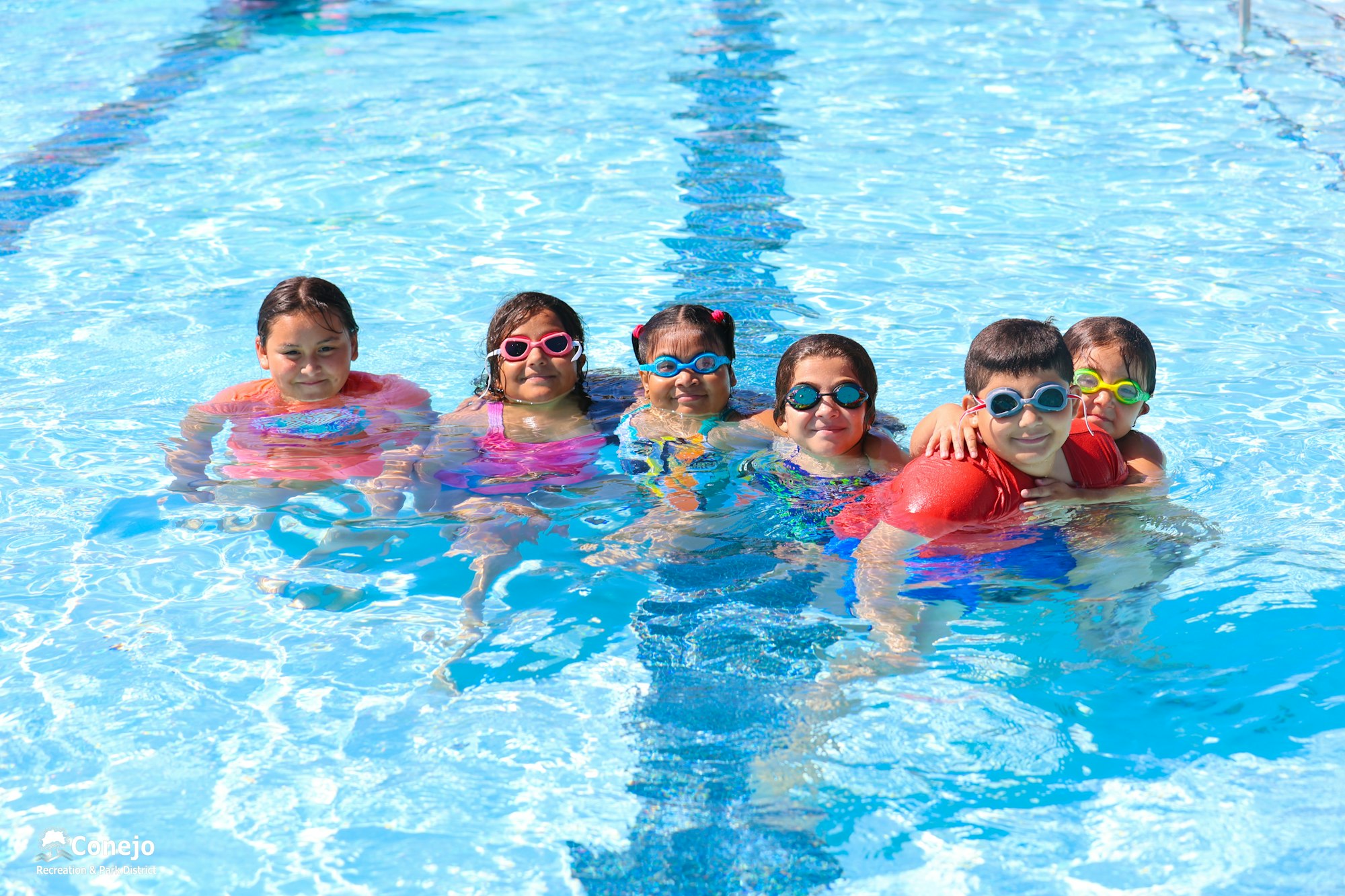 Seven children in a pool wearing colorful swimsuits and goggles.