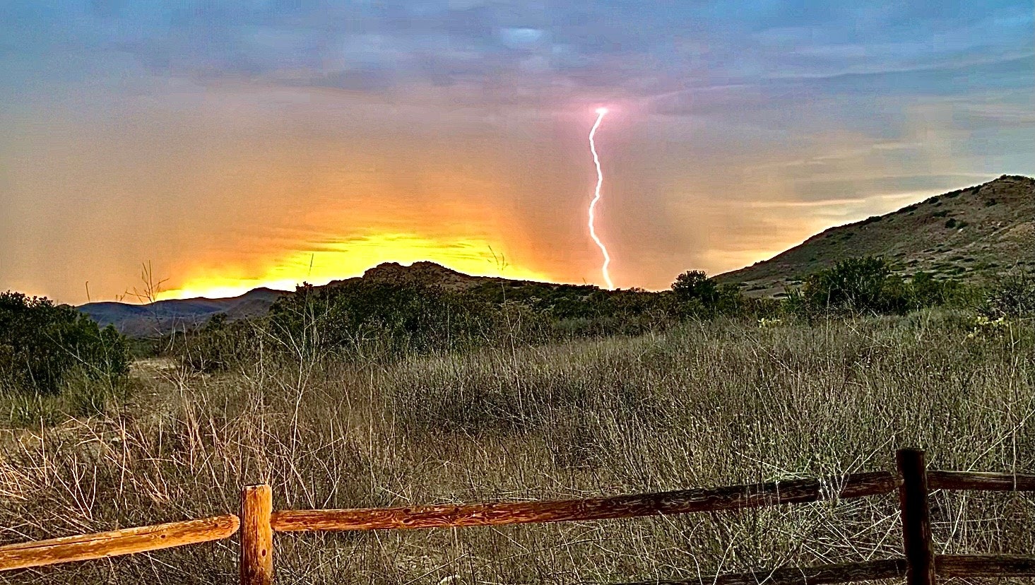 A lightning bolt strikes during a sunset over hilly terrain, with a wooden fence in the foreground.