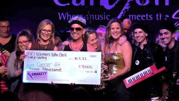 A group holding a $7,000 donation check for "Cancer Fit" at a music event, with people smiling and instruments in the background.