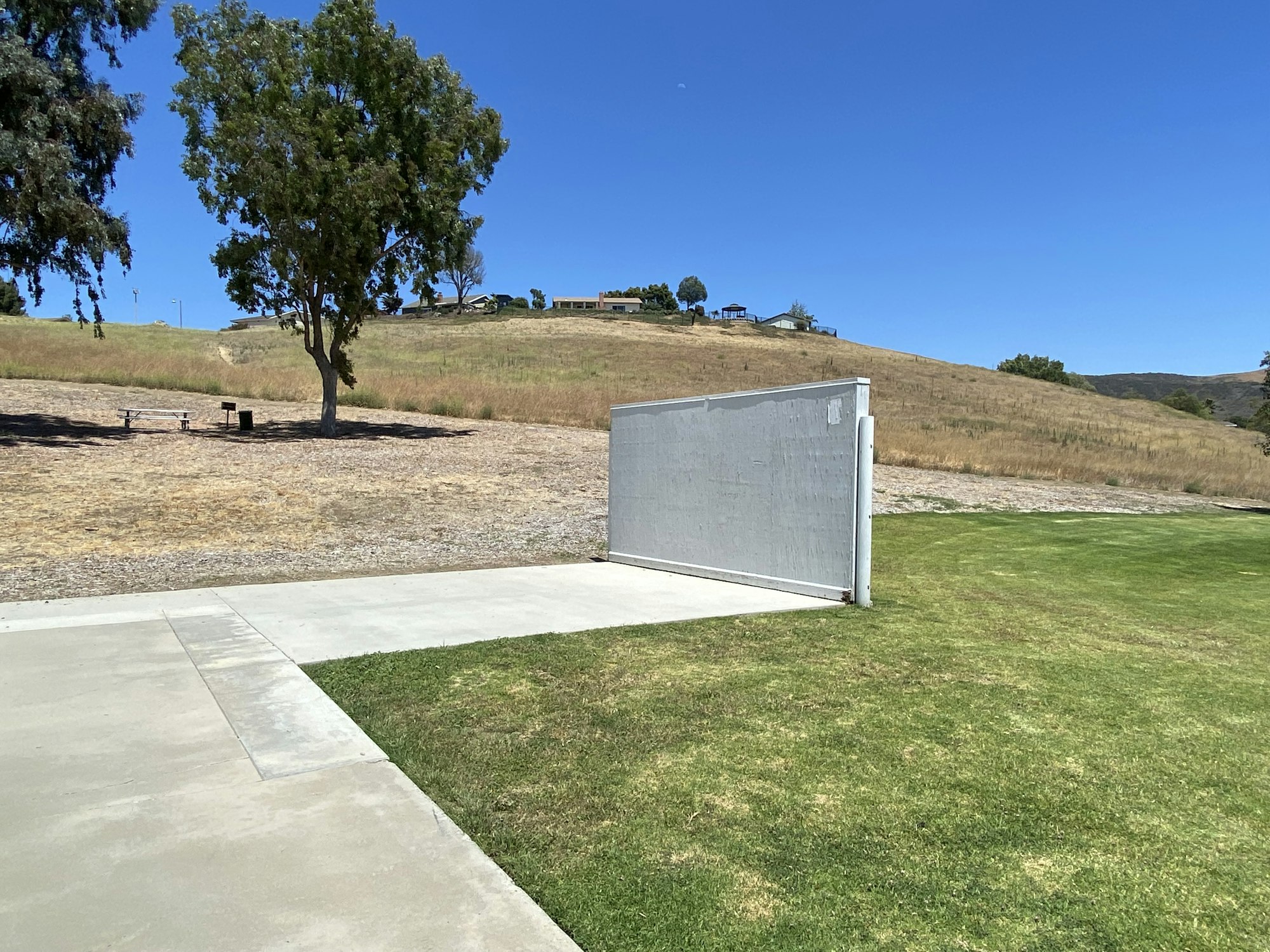 A lone wall in a park with a tree, picnic table, and rolling hills under a clear blue sky.