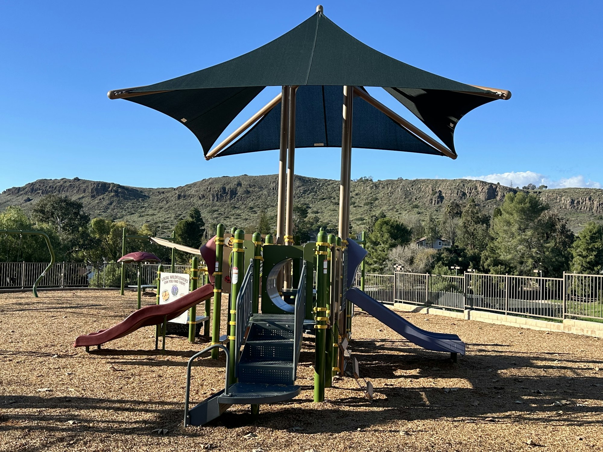 Playground with slides and a large canopy, fences, and trees under a clear blue sky.