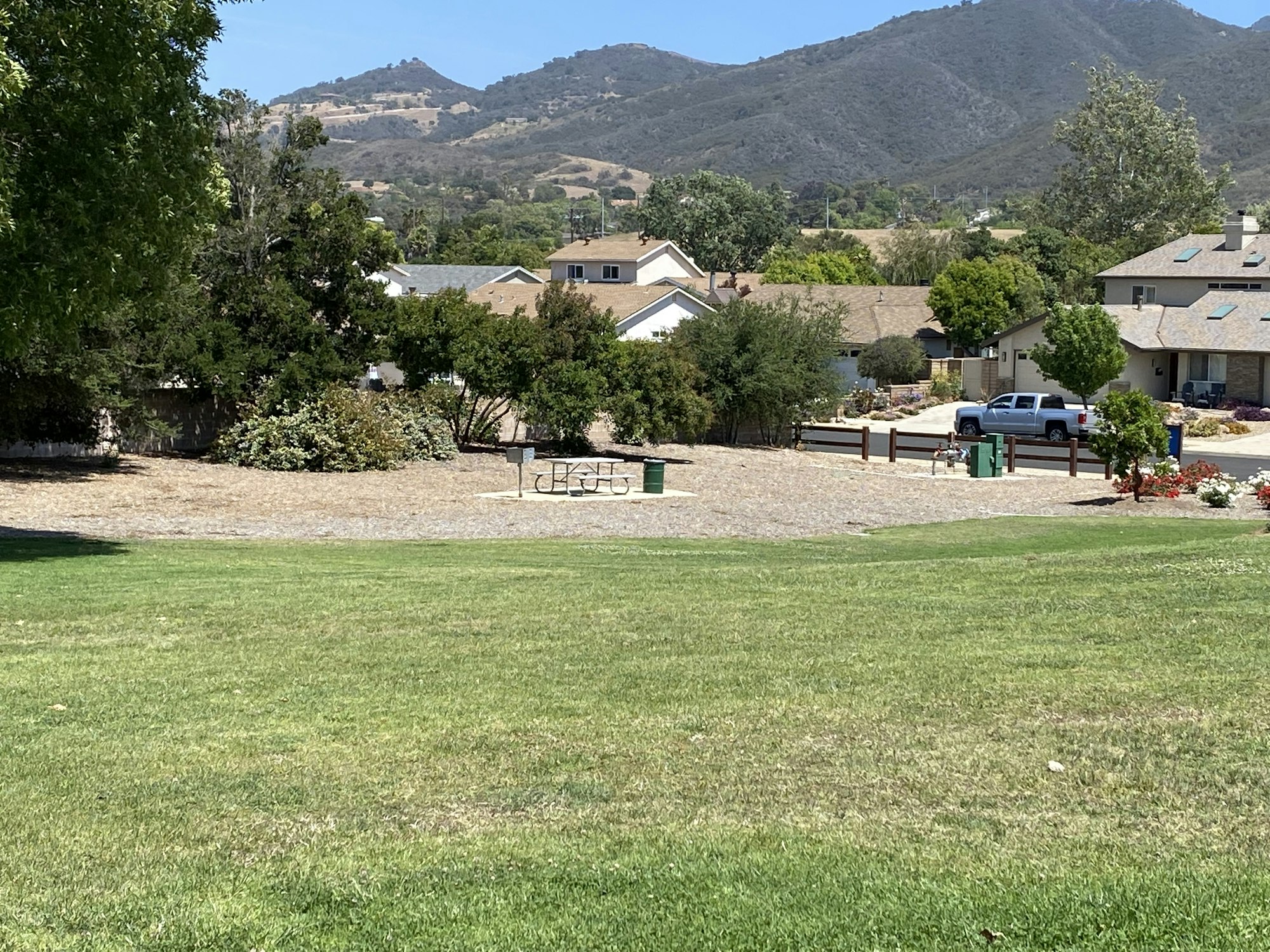 A green grassy area with distant residential homes, hills, trees, a picnic table, and a parked vehicle.