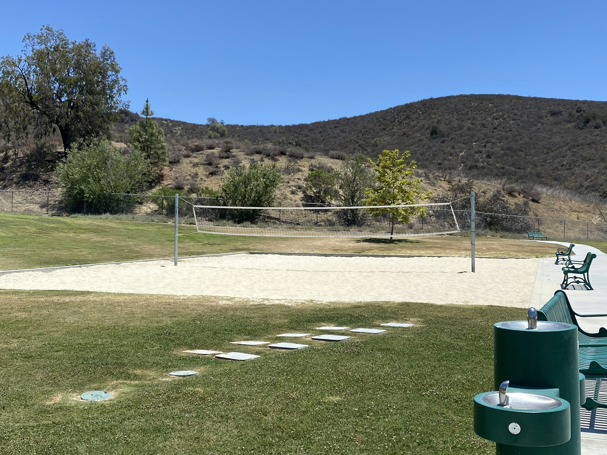 Outdoor volleyball court with sand, surrounded by grass, benches, and water fountains, set against a hill and clear blue sky.
