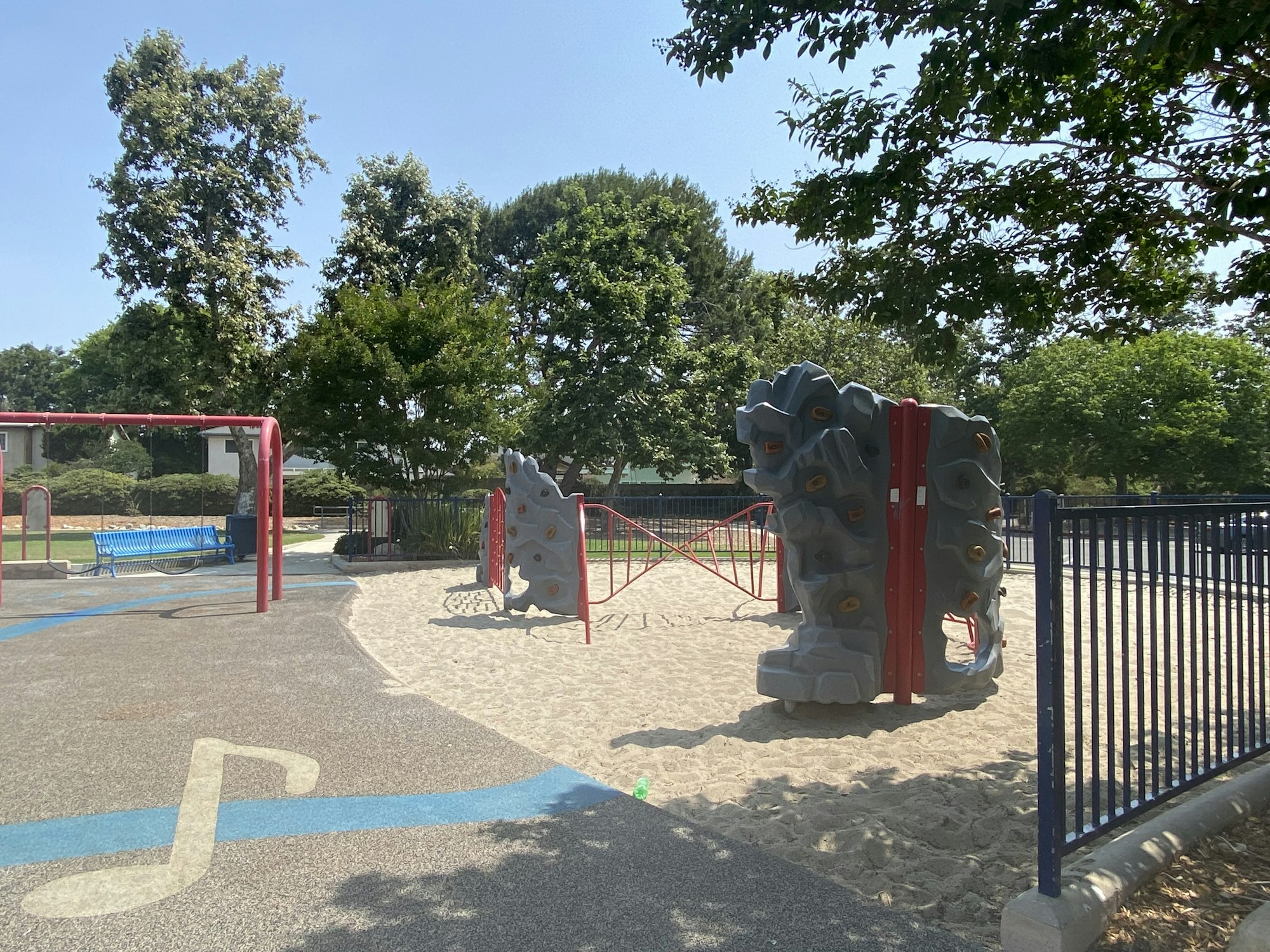 Playground with climbing structures, swings, sand, music note design, trees, and a blue bench.