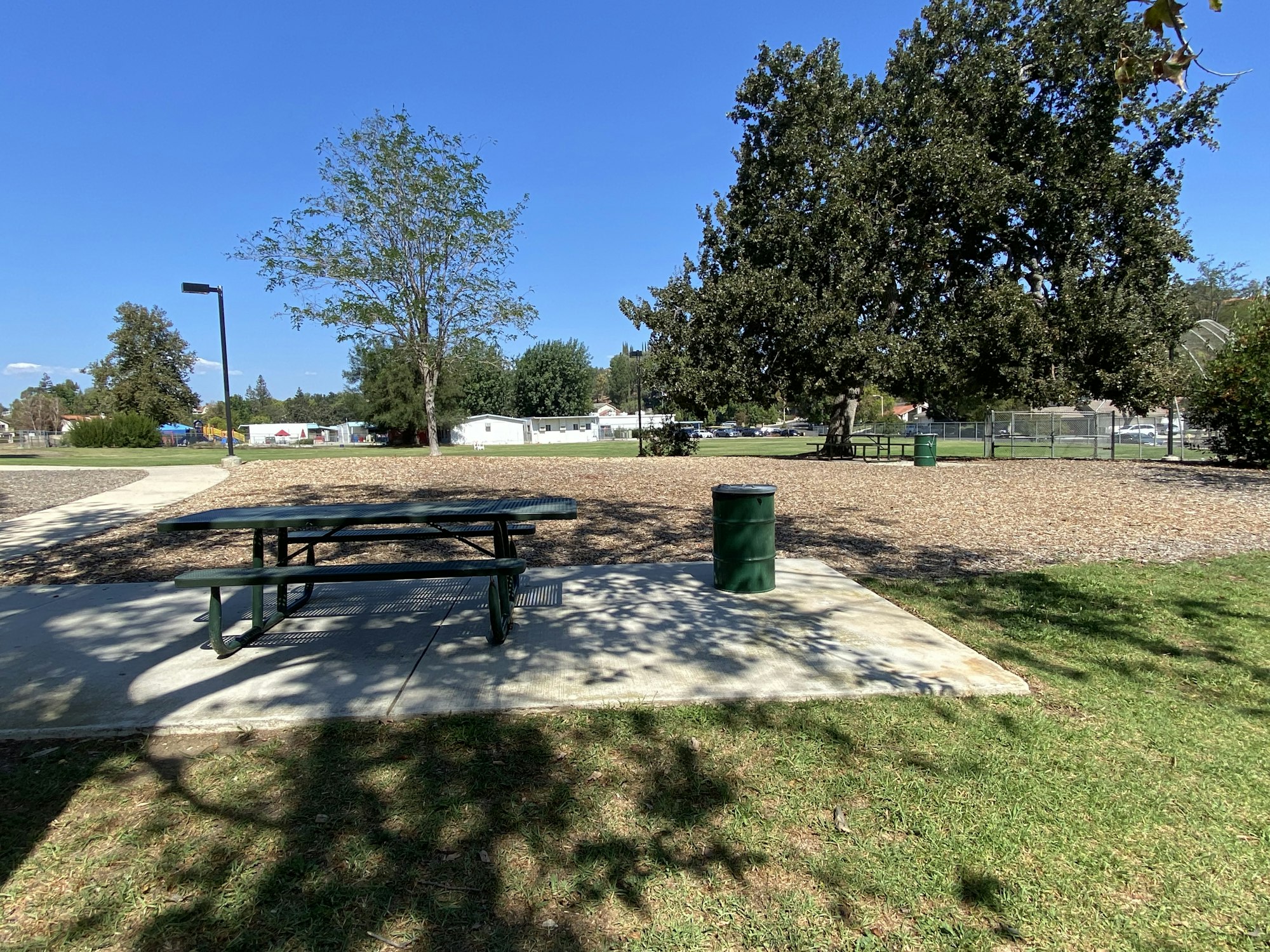 A park with picnic tables, trash bins, trees, and a playground in the background under a clear blue sky.