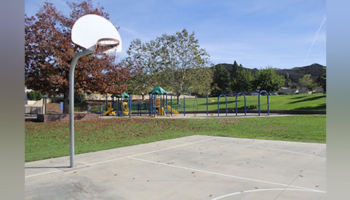 A basketball court next to a playground with trees and grass under a clear sky.