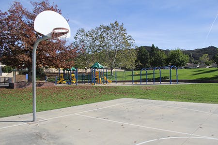 A basketball court next to a playground with trees and grass under a clear sky.