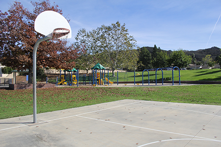 A basketball court next to a playground with trees and grass under a clear sky.