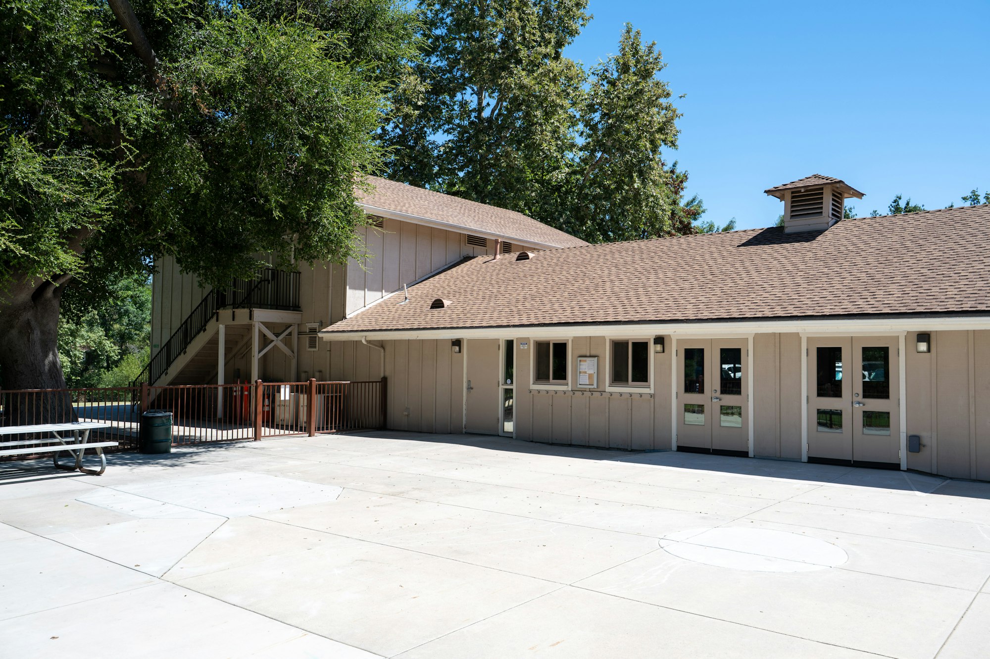 A building with a brown roof, trees nearby, exterior doors and windows, and a concrete area with seating.