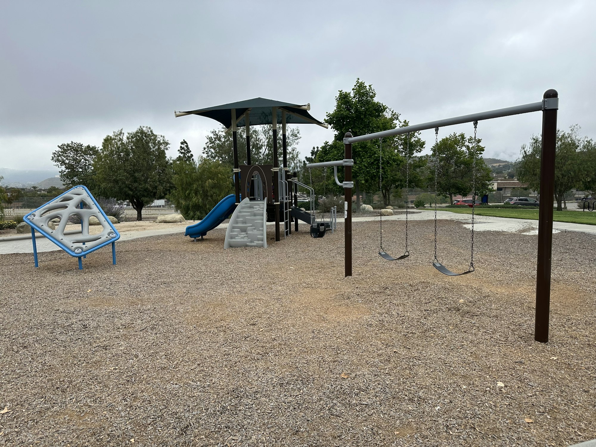 A playground featuring a slide, climbing structure, swings, and a gravel surface under a cloudy sky.
