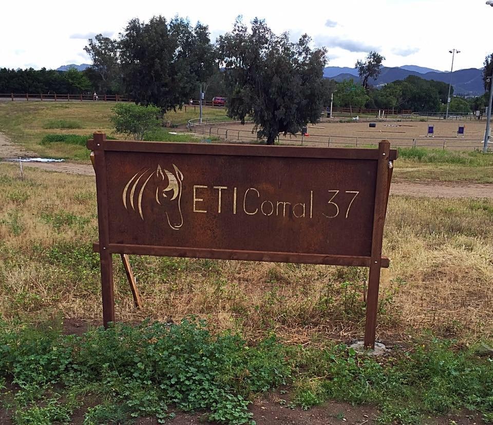 Metal sign reading "ETI Corral 37" with a horse head design, in a grassy field with trees and mountains in the background.