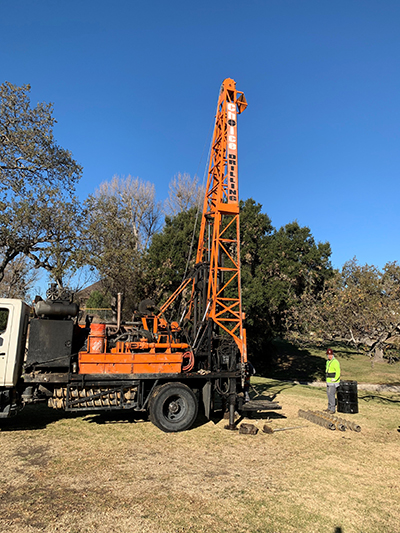 A drilling rig on a grassy area with a person in a high-visibility jacket nearby, under a clear blue sky.