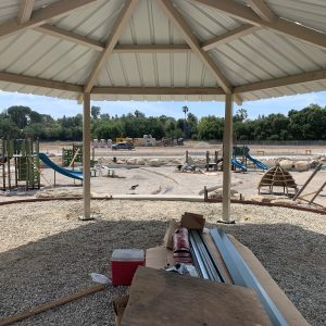 A gazebo overlooks a playground under construction with slides, equipment, and scattered building materials.