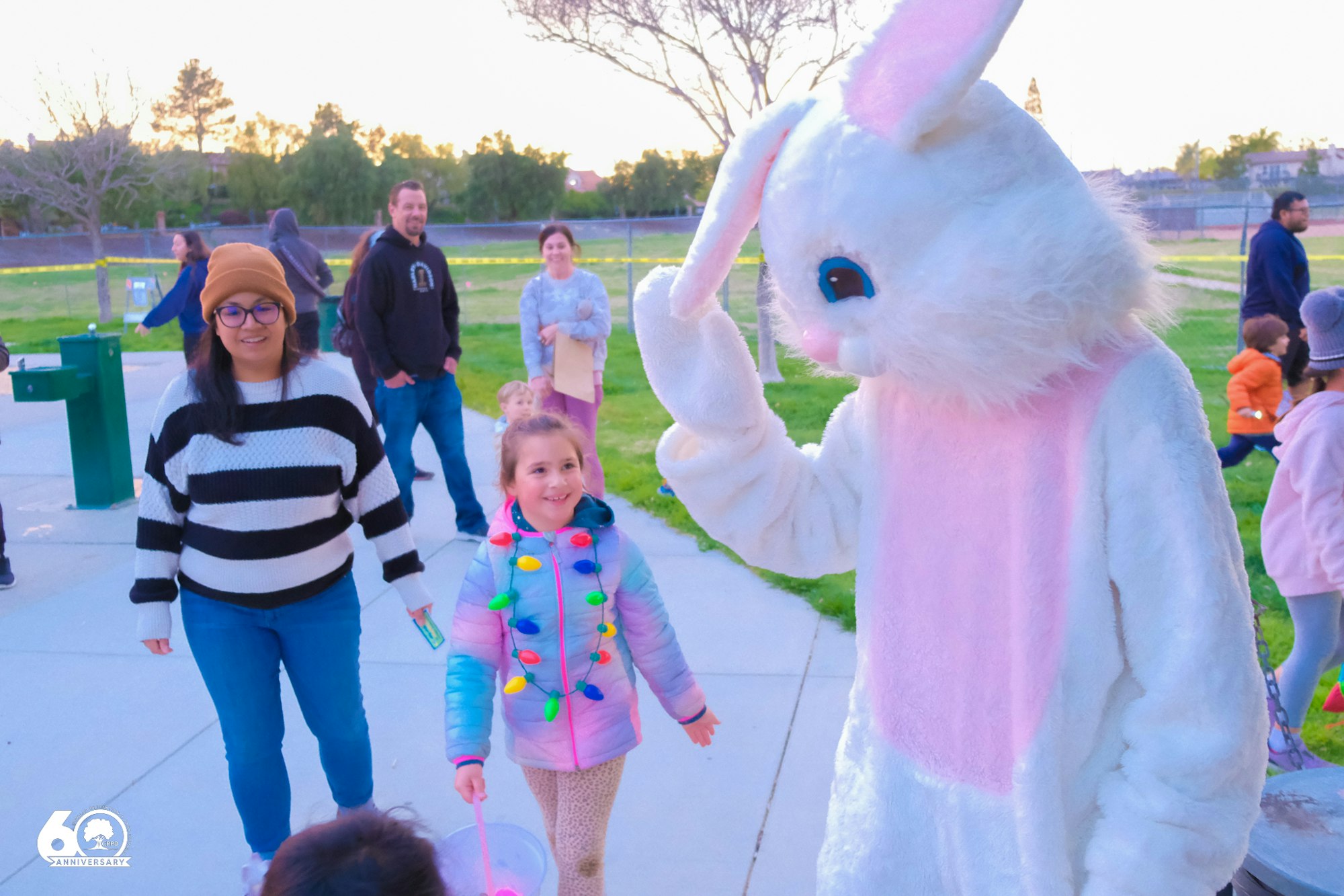 Person in Easter Bunny costume greeting children at a park; people nearby enjoying the event.