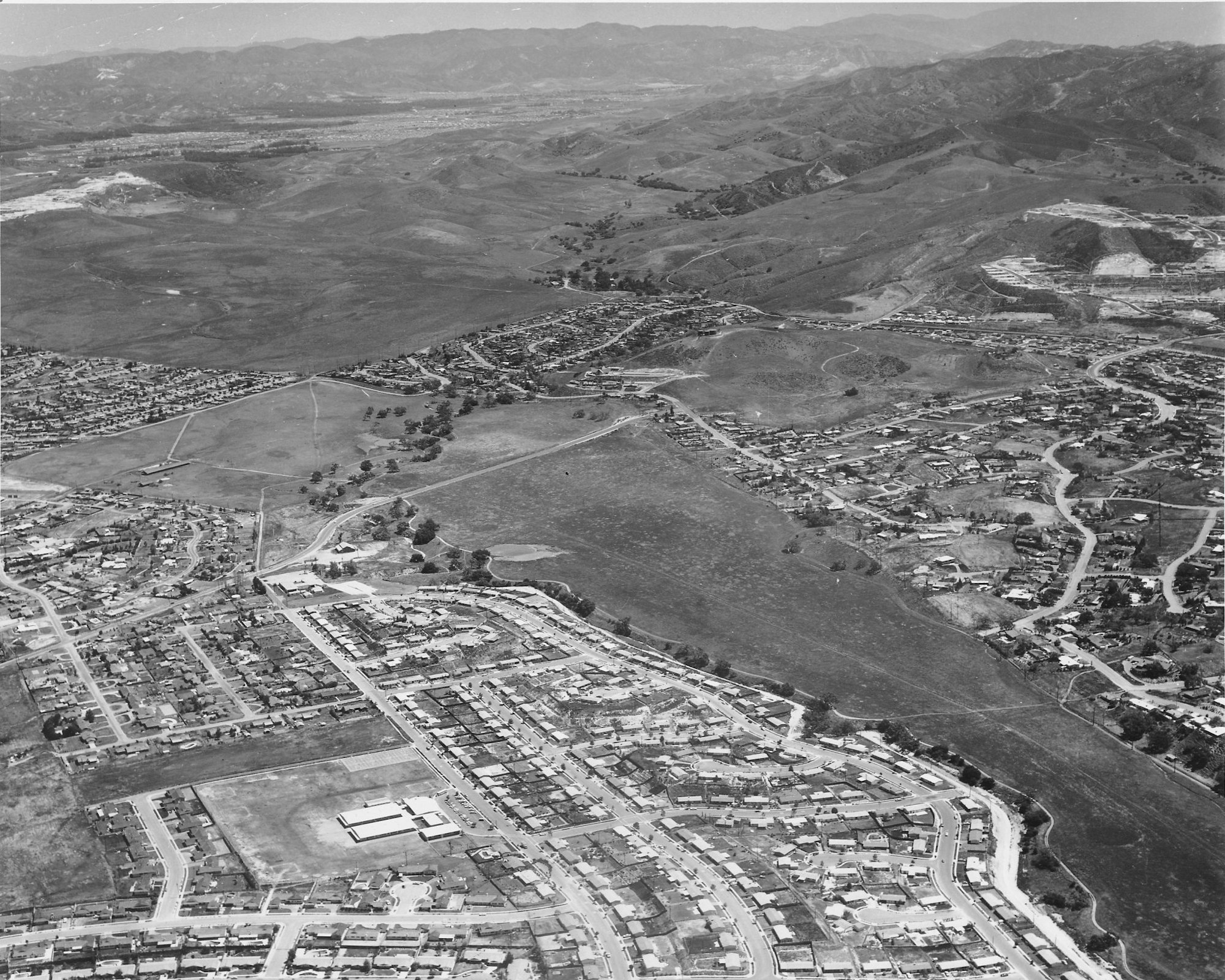 Aerial view of a suburban neighborhood with houses, roads, open fields, and mountains in the background.