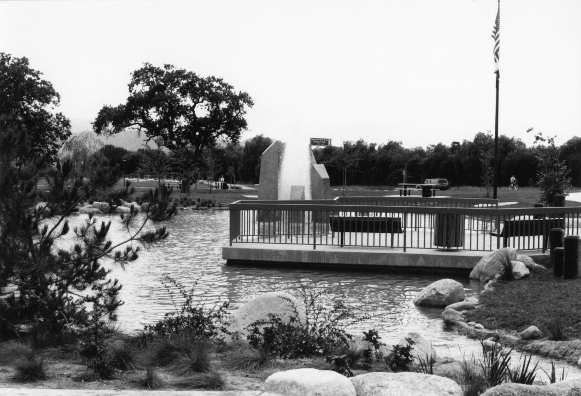 Park scene with a fountain, trees, a flagpole, and a pond. There's a person cycling and a car in the background.