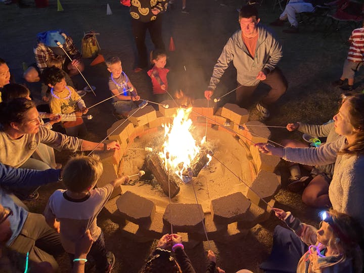 People roasting marshmallows over a campfire at night, with kids and adults gathered around.