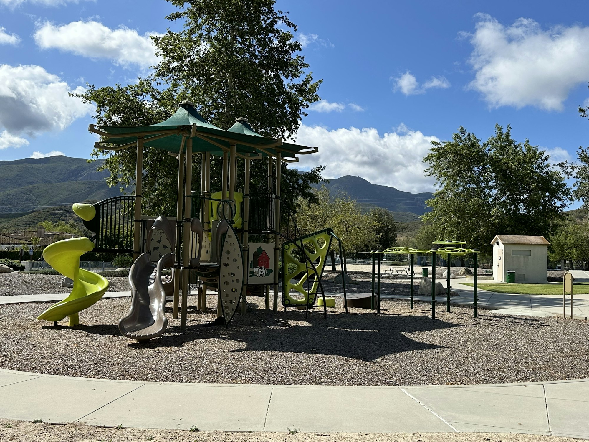 A playground with green and gray slides, climbing structures, and a restroom, set against a backdrop of mountains and blue skies.