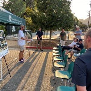 A man speaks to a small outdoor gathering, with visual displays and chairs set up under a canopy.