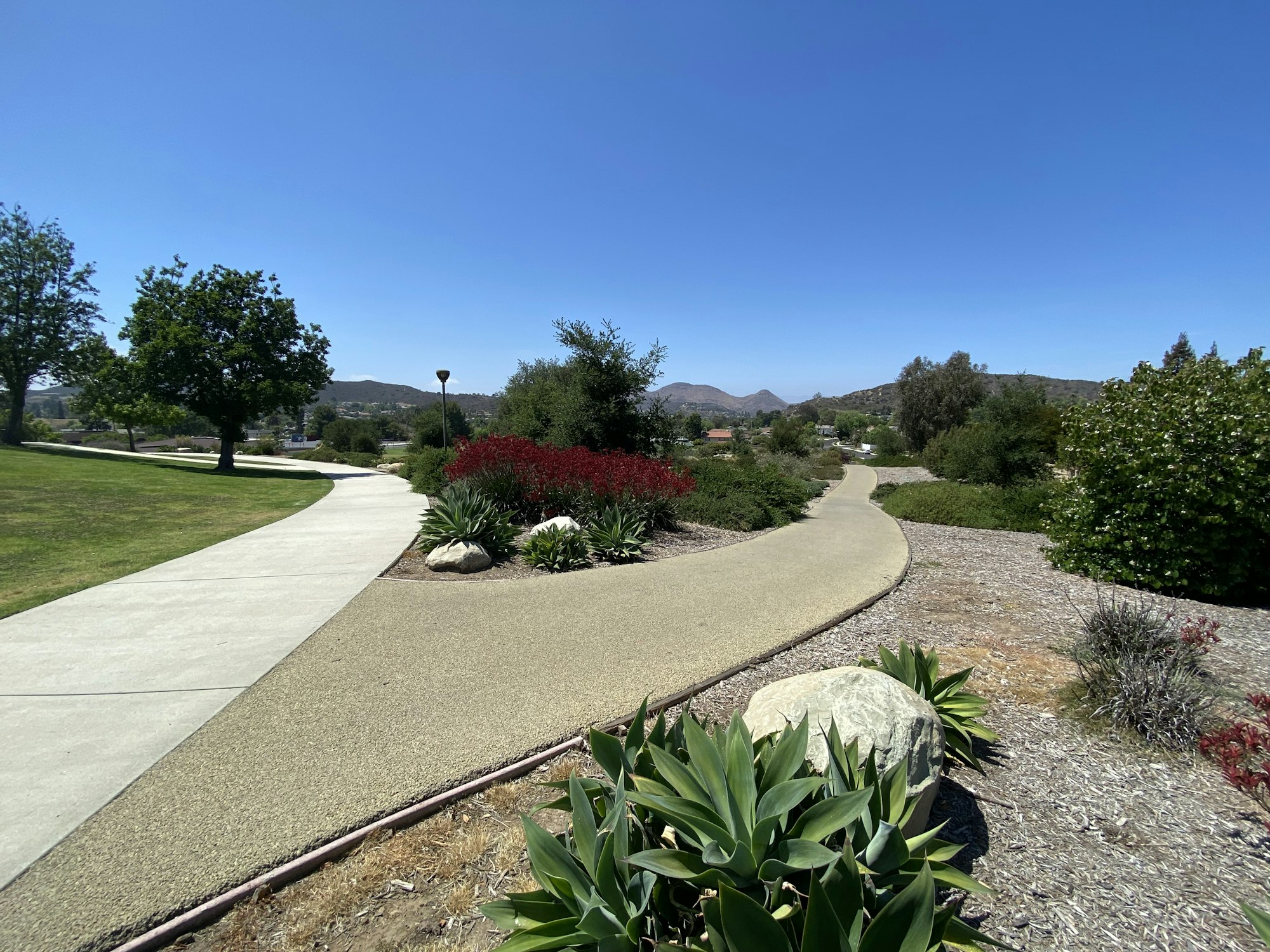 A scenic park with paved paths, green grass, shrubs, and distant mountains under a clear blue sky.