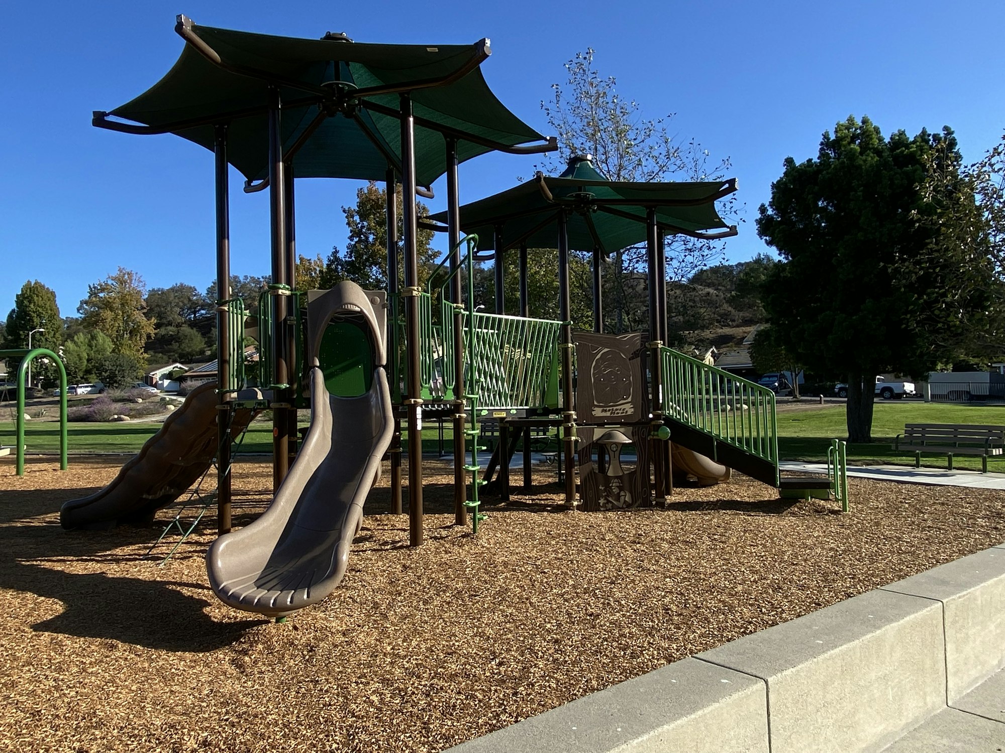 Playground with slides and climbing structures, surrounded by wood chips, trees, and a clear blue sky.