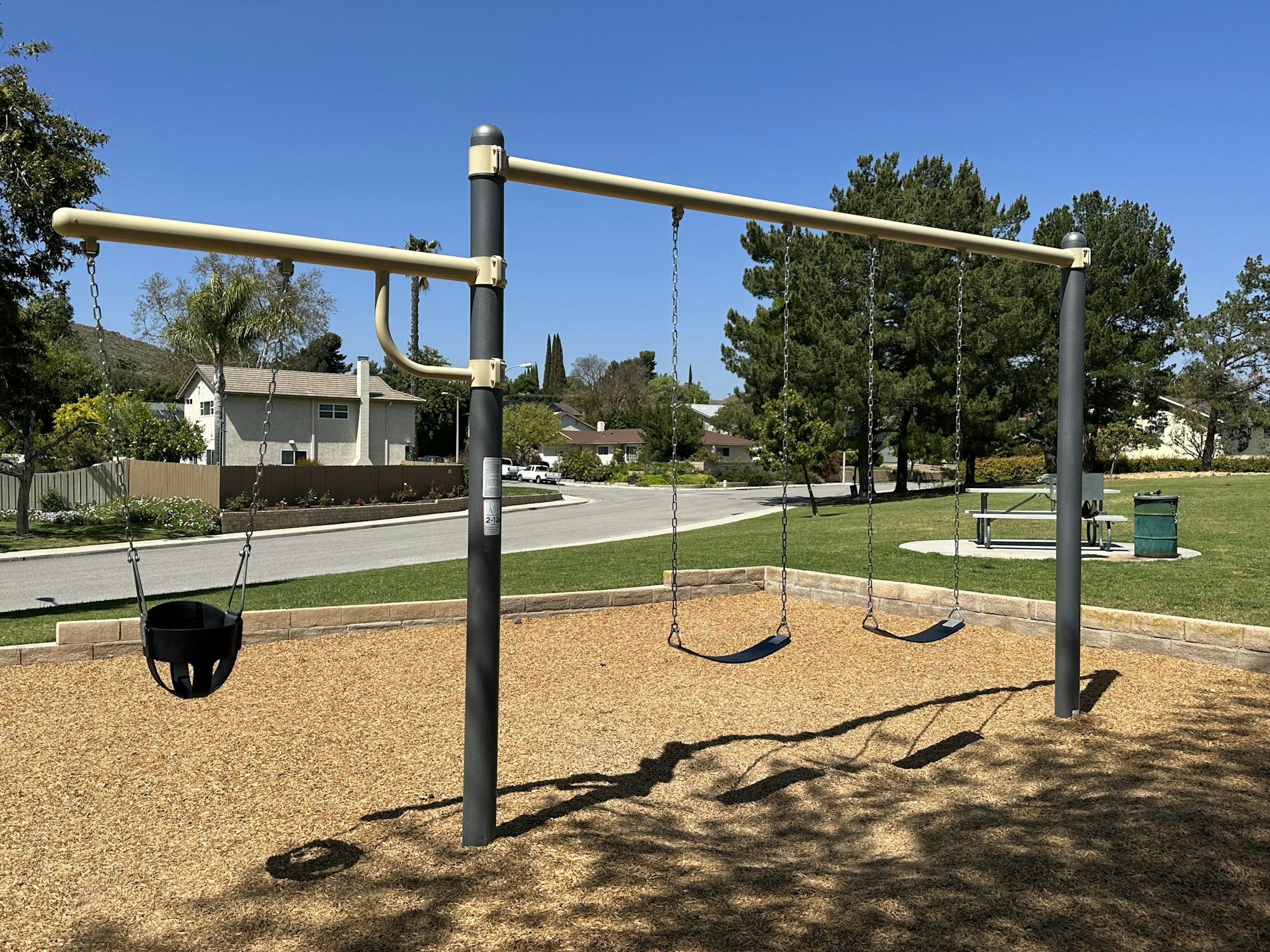 Playground with two swings and a toddler swing, trees, houses, and a picnic area in the background.