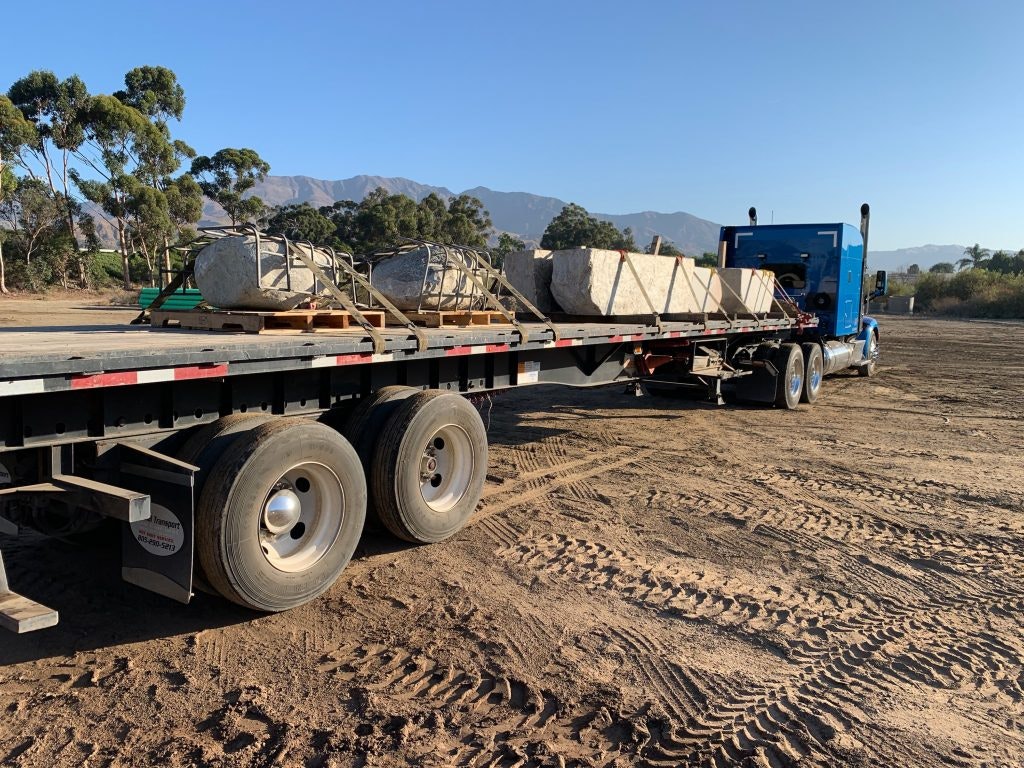 A blue semi-truck with a flatbed trailer carrying large boulders on a dirt lot, trees and mountains in the background.