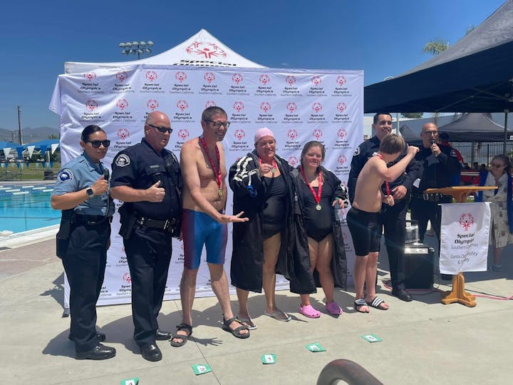 Group celebrates at Special Olympics Southern California event, posing with medals and two officers near a pool.