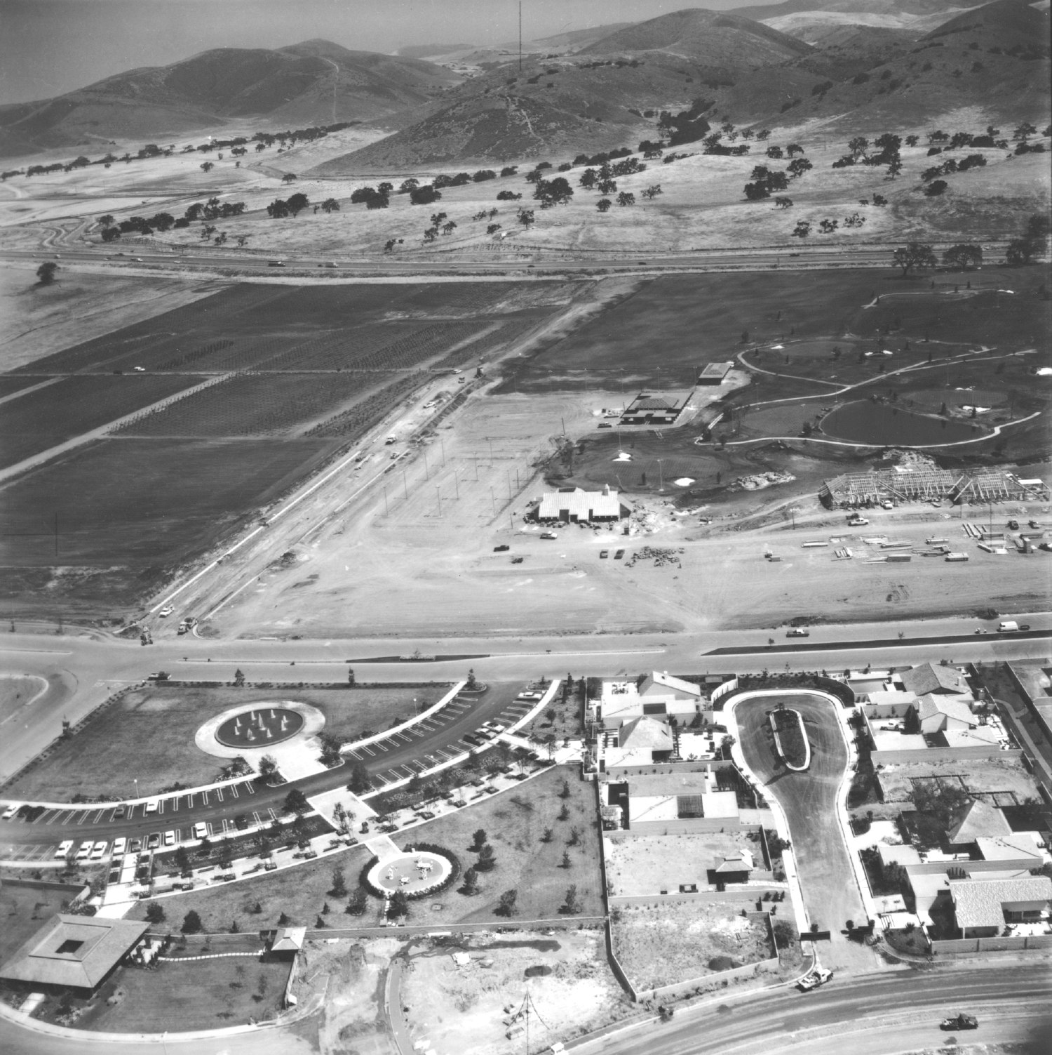 Aerial view of a developing suburban area and farmland near hills, featuring roads, parking lots, and residential houses.