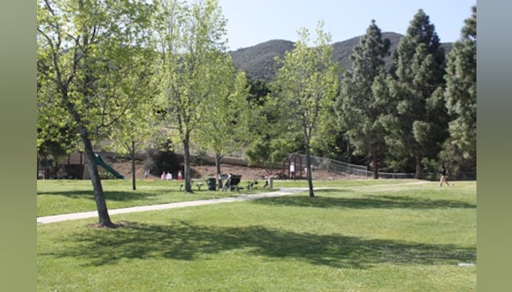 A park with trees, a grassy area, a playground, people in the distance, and hills in the background.