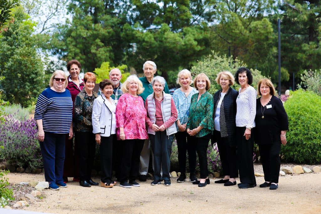 A group of 13 people posing outdoors, surrounded by greenery and flowers.